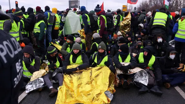 Протестующие пытаются заблокировать дорогу к месту проведения конференции АдГ. Люди в зеленых светоотражающих жилетах сидят на асфальте, укрывшись одеялами из фольги, некоторые из них стоят, слева в кадре виден полицейский в форме. У протестующих на заднем фоне флаги, в том числе один флаг Палестины.