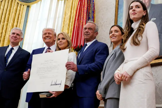 Кеннеди позирует со свидетельством, подтверждающим его должность. Рядом его жена, Трамп и другие люди.