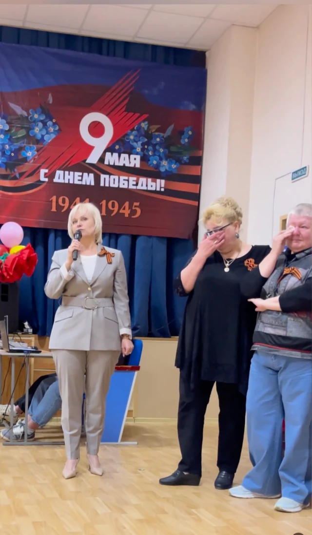 Женщина в брючном костюме с микрофоном, две другие плачут