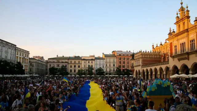 Большая толпа людей на площади, окруженной зданиями с трех сторон. В центре толпы развевается большой сине-желтый украинский флаг. 24 августа 2023 г.