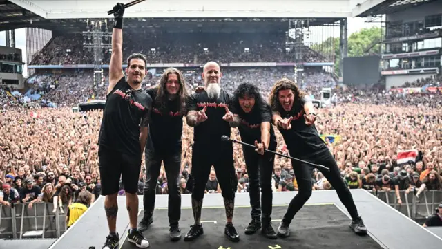 Пять участников группы Anthrax позируют на сцене перед огромной толпой зрителей на стадионе.