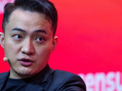 Джастин Сан в черном пиджаке и черной футболке, с маленьким бежевым микрофоном и наушником, на фоне красной стены