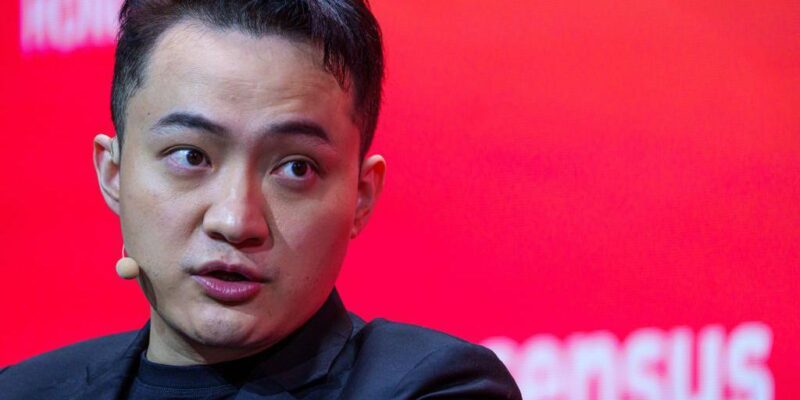 Джастин Сан в черном пиджаке и черной футболке, с маленьким бежевым микрофоном и наушником, на фоне красной стены