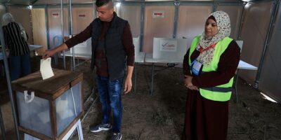 В городе Дейр-эль-Балах в центре сектора Газа, единственном городе, где организованы эти выборы, соорудили временные участки для голосования. Мужчина опускает бюллетень в урну, за этим наблюдает женщина в светоотражающем жилете и платке