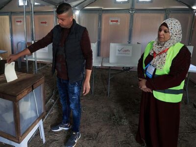 В городе Дейр-эль-Балах в центре сектора Газа, единственном городе, где организованы эти выборы, соорудили временные участки для голосования. Мужчина опускает бюллетень в урну, за этим наблюдает женщина в светоотражающем жилете и платке