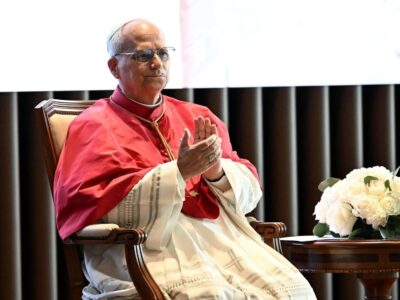 Папа Лев в белой сутане и красной моццетте сидит в кресле и хлопает в ладоши, на столике рядом с ним букет белых цветов