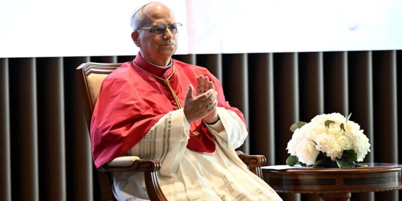 Папа Лев в белой сутане и красной моццетте сидит в кресле и хлопает в ладоши, на столике рядом с ним букет белых цветов