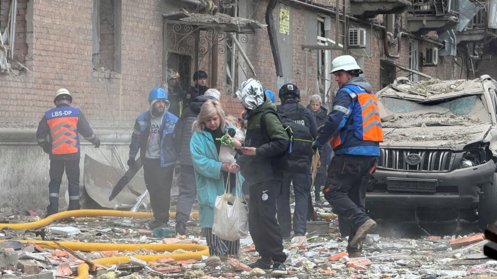 Женщина с собачкой в руках выходит из дома, на земле — обломки кирпичей, рядом с ней несколько человек в форме спасателей.