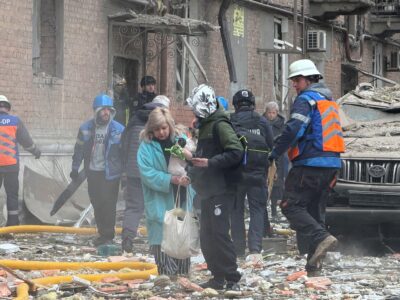 Женщина с собачкой в руках выходит из дома, на земле — обломки кирпичей, рядом с ней несколько человек в форме спасателей.