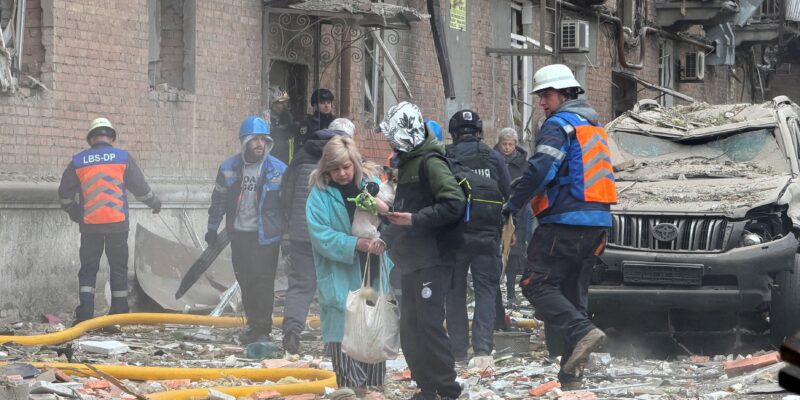 Женщина с собачкой в руках выходит из дома, на земле — обломки кирпичей, рядом с ней несколько человек в форме спасателей.