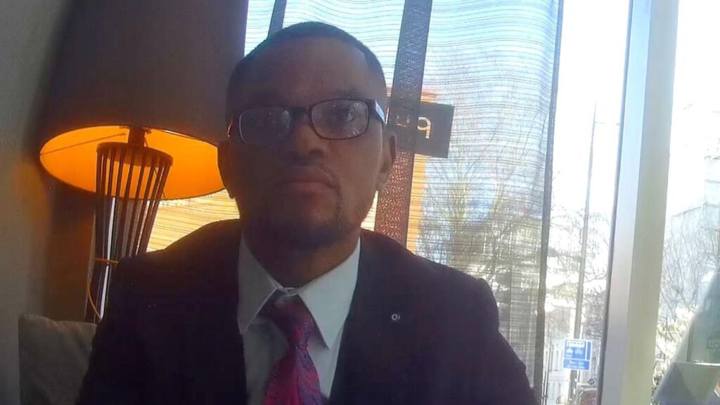 Фейковый юридический консультант, секретная съемка: темнокожий мужчина в очках, в пиджаке, белой рубашке и галстуке, на фоне окна и зажженной настольной лампы в лобби отеля.