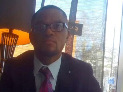 Фейковый юридический консультант, секретная съемка: темнокожий мужчина в очках, в пиджаке, белой рубашке и галстуке, на фоне окна и зажженной настольной лампы в лобби отеля.