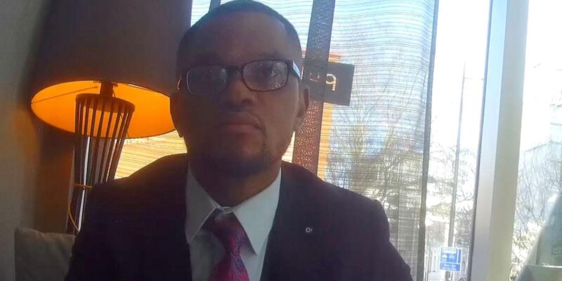 Фейковый юридический консультант, секретная съемка: темнокожий мужчина в очках, в пиджаке, белой рубашке и галстуке, на фоне окна и зажженной настольной лампы в лобби отеля.