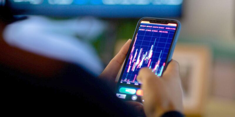 Man looking at trading app on his smart phone from his home office. He is being photographed from behind, so only his hands on his phone are visible in hard focus with the back of his shoulders and his office screens visible in soft focus.