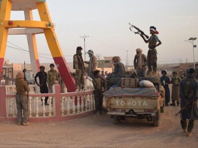 Группа боевиков в камуфляжной форме стоит вокруг пикапа на пыльной дороге. Одним из них стоит на автомобиле и держит в руках автомат. Слева на снимке видны коричнево-желтые ноги скульптуры.