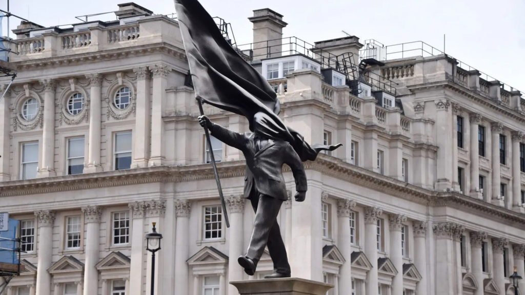 Скульптура: мужчина в костюме шагает вперед с постамента, одной рукой держа флаг, и этот же флаг закрывает его лицо.