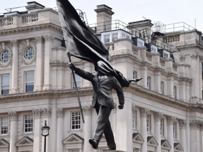 Скульптура: мужчина в костюме шагает вперед с постамента, одной рукой держа флаг, и этот же флаг закрывает его лицо.