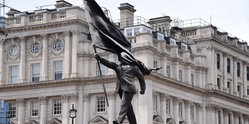 Скульптура: мужчина в костюме шагает вперед с постамента, одной рукой держа флаг, и этот же флаг закрывает его лицо.