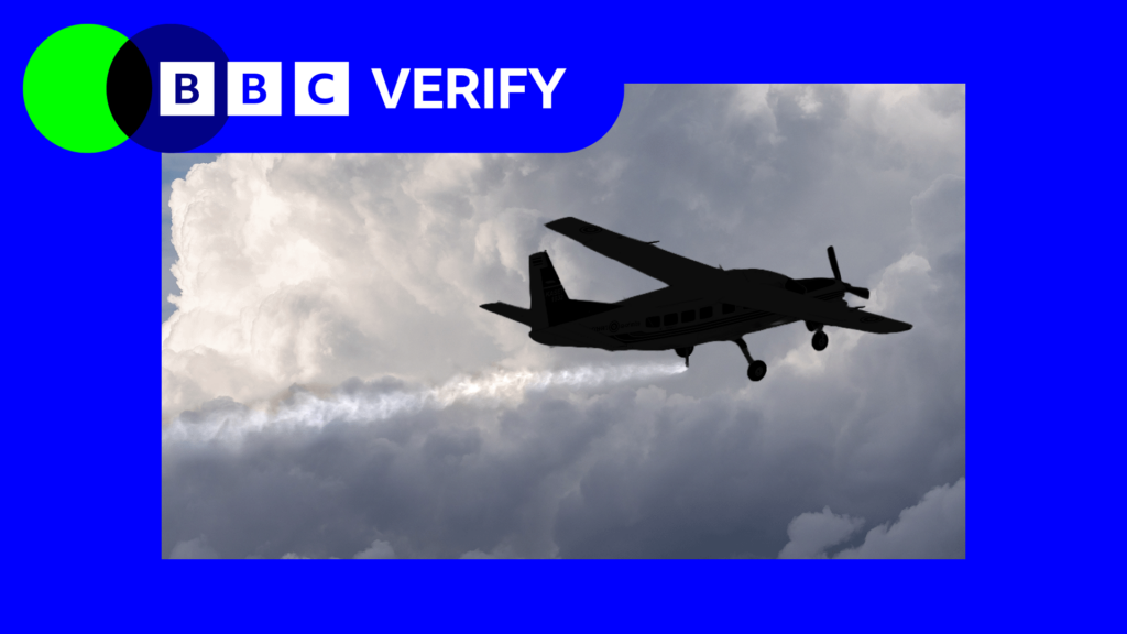 Небольшой самолет летит на фоне плотных серых облаков, распыляя мелкие частицы, которые образуют длинный белый след.