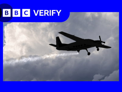 Небольшой самолет летит на фоне плотных серых облаков, распыляя мелкие частицы, которые образуют длинный белый след.