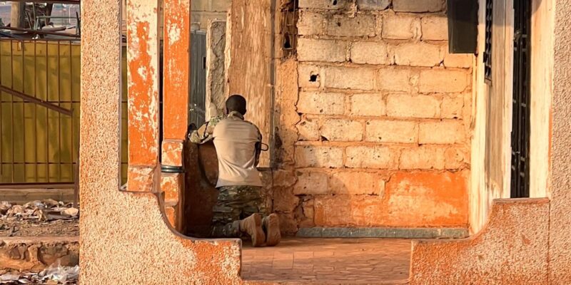 Мужчина в камуфляже с оружием стоит на коленях в кирпичном здании и наблюдает в окно