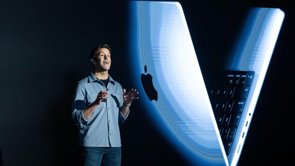 Джон Тернус на черном фоне, за спиной большое изображение макбука