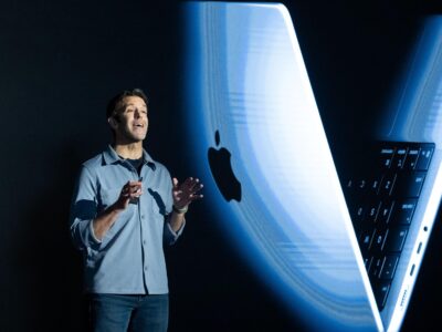 Джон Тернус на черном фоне, за спиной большое изображение макбука