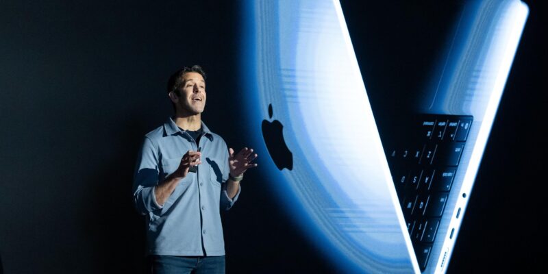 Джон Тернус на черном фоне, за спиной большое изображение макбука