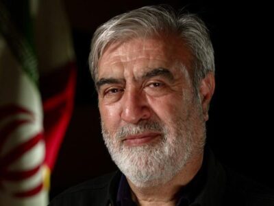 Мужчина с седой бородой и седыми волосами смотрит в камеру. На нем черная рубашка, а за его спиной висит иранский флаг.