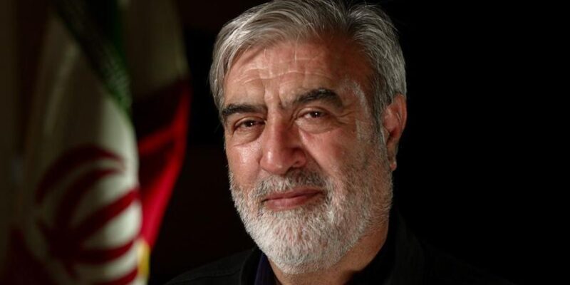 Мужчина с седой бородой и седыми волосами смотрит в камеру. На нем черная рубашка, а за его спиной висит иранский флаг.