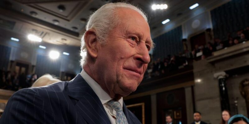 Король Карл в синем костюме в тонкую полоску, белой рубашке и светло-голубом галстуке.