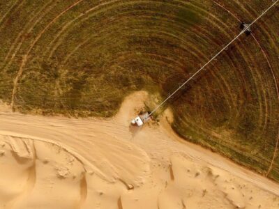Вид с высоты на круглое зеленое сельскохозяйственное поле, граничащее с пустынным песком, с длинным трубопроводом, идущим от небольшого белого сооружения к зеленой зоне.