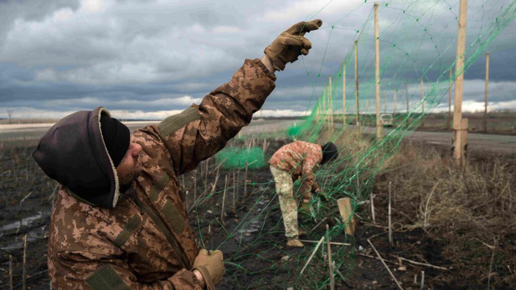 Двое украинских военных натягивают антидроновую сетку на столбах вдоль дороги