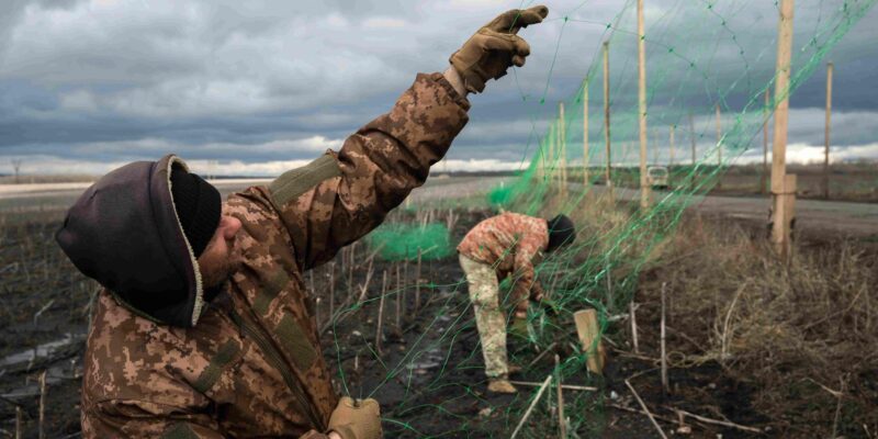 Двое украинских военных натягивают антидроновую сетку на столбах вдоль дороги
