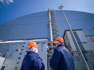 Двое работников ЧАЭС в оранжевых шлемах стоят у купола, защищающего саркофаг