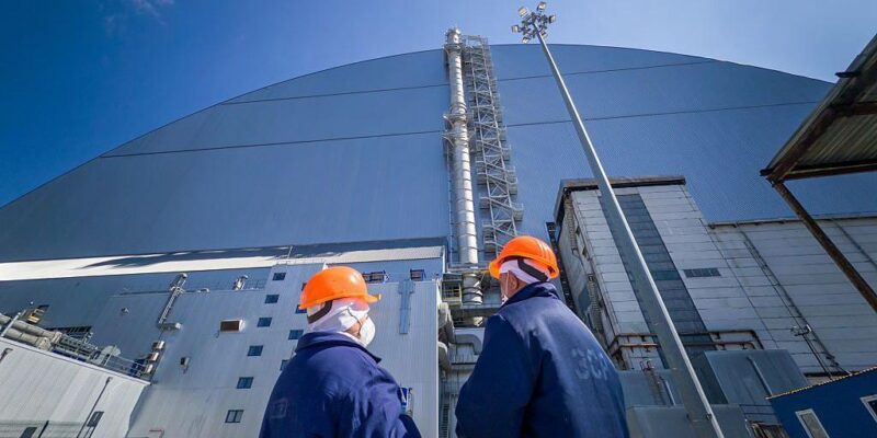 Двое работников ЧАЭС в оранжевых шлемах стоят у купола, защищающего саркофаг