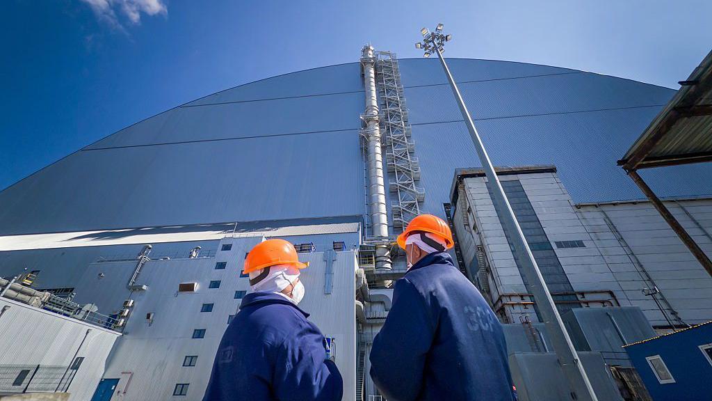 Двое работников ЧАЭС в оранжевых шлемах стоят у купола, защищающего саркофаг