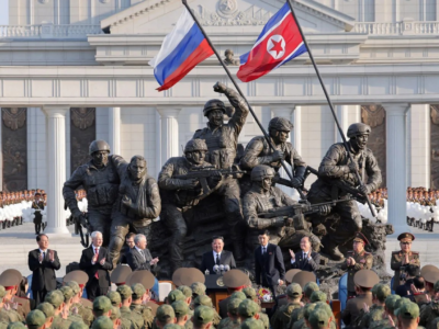 Монумент в виде скульптурной группы: несколько военных держат оружие, один эвакуирует раненого, двое держат флаги, российский и северокорейский. Перед памятником стоят чиновники и военные, на заднем плане здания с колоннами, возле которого выстроились в ряды военные с белыми шарами.