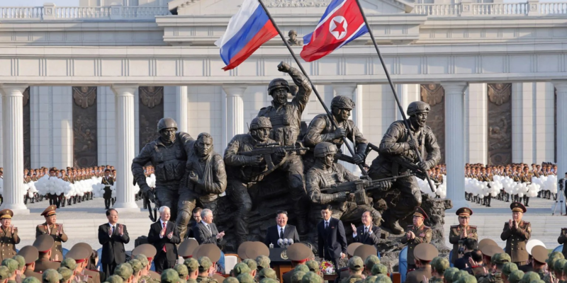 Монумент в виде скульптурной группы: несколько военных держат оружие, один эвакуирует раненого, двое держат флаги, российский и северокорейский. Перед памятником стоят чиновники и военные, на заднем плане здания с колоннами, возле которого выстроились в ряды военные с белыми шарами.
