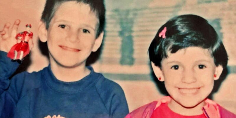 A boy in a blue top and a girl in a pink top, both with dark hair, smile at the camera - the boy is holding up a toy figurine