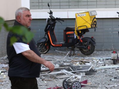 «Если выход завалит — все!» Почему Подмосковье оказалось не готово к атакам беспилотников 4 На заданем плане стена многоквартирного дома. Перед ней припаркован фиолетовый электросамокат. Рядом стоит курьерский электросамокат с желтым коробом на багажнике. Земля перед домам усыпано осколками стекла и обломками выбитых окон. На переднем плане слева направо идет мужчина.
