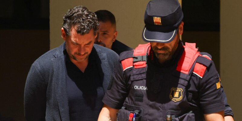 Мужчина в черной кофте с руками за спиной идет, понурив голову, рядом с полицейским в бронежилете.