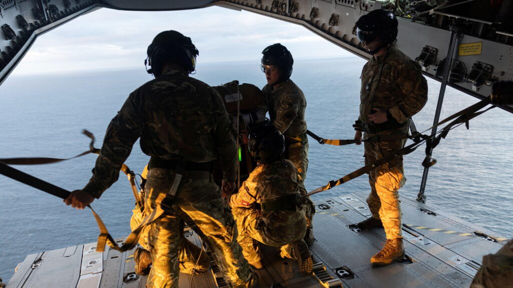 оеннослужащие на краю самолёта в полевой форме; под ними — водная поверхность, они готовят предметы для сброса с воздуха.