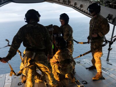 оеннослужащие на краю самолёта в полевой форме; под ними — водная поверхность, они готовят предметы для сброса с воздуха.