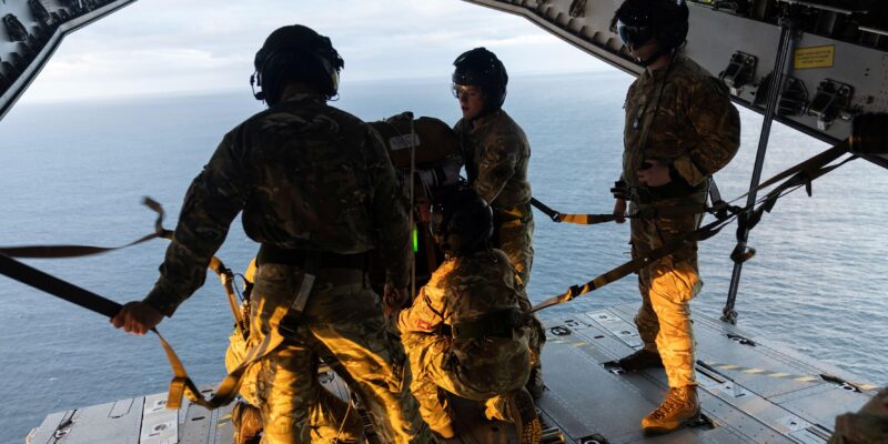 оеннослужащие на краю самолёта в полевой форме; под ними — водная поверхность, они готовят предметы для сброса с воздуха.
