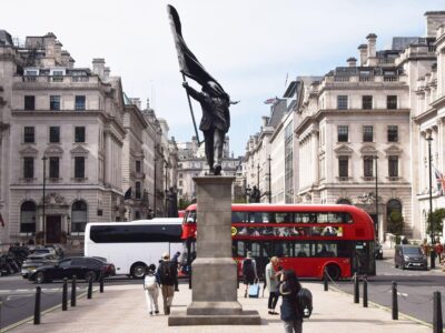 A grey statue of a man wearing a suit, walking forward off a plinth, carrying a flag that covers his face. A red London bus can be seen in th bcakground