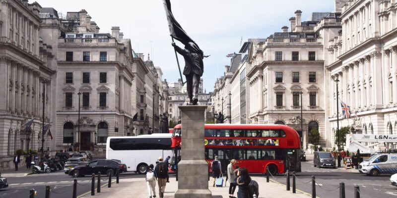 A grey statue of a man wearing a suit, walking forward off a plinth, carrying a flag that covers his face. A red London bus can be seen in th bcakground