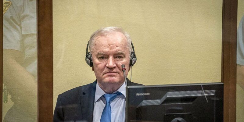 Ратко Младич в костюме с галстуком сидит за стеклом, на нем наушники, перед ним микрофон и монитор.