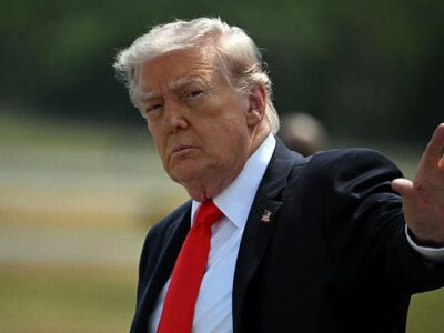 Дональд Трамп, одетый в костюм, поднимает руку в сторону камеры.