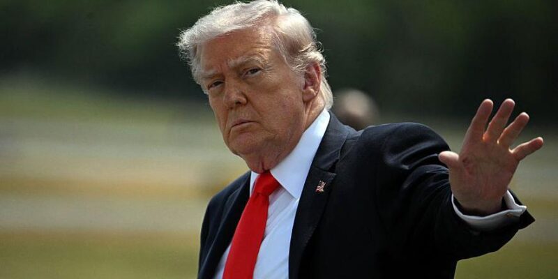 Дональд Трамп, одетый в костюм, поднимает руку в сторону камеры.